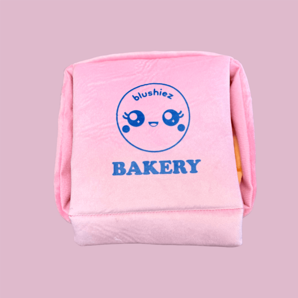 Pink plush bakery box dog toy with blue face logo and 'BAKERY' text