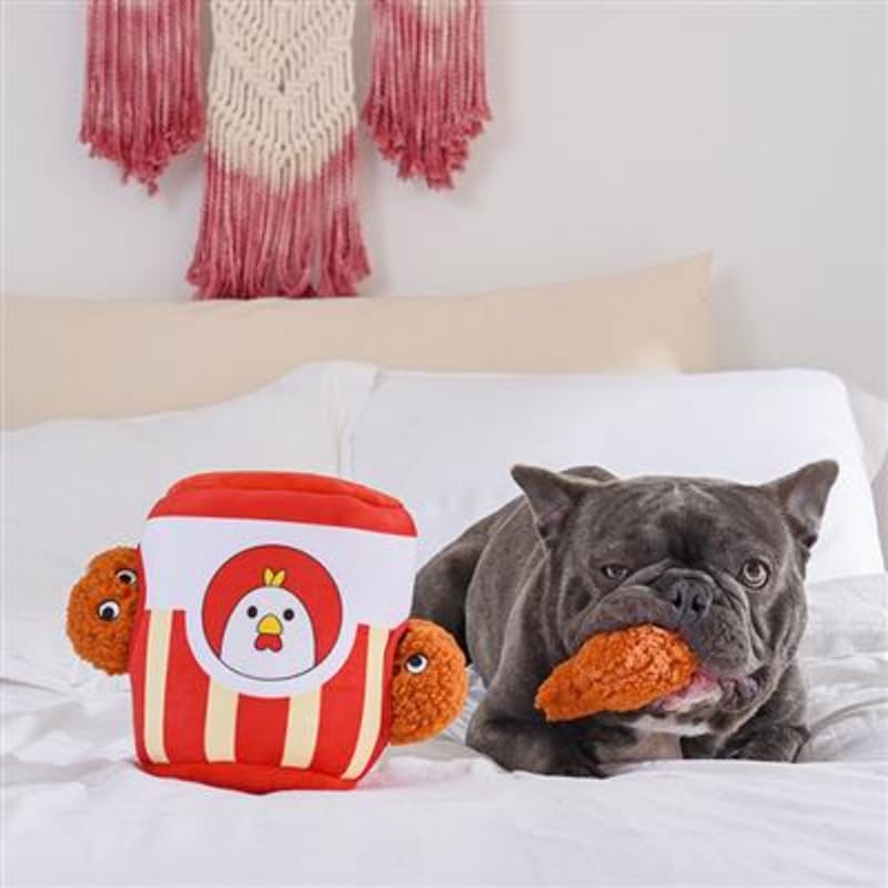 Interactive plush dog toy on bed with lying dog; red-and-white drumstick can and plush pieces