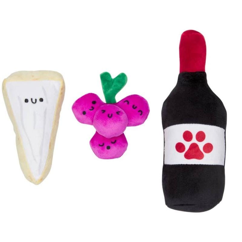 Close-up: three plush dog toys—cream bone, purple critter with green antlers, black paw bottle