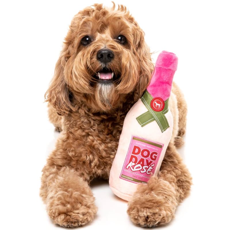 Plush bottle-shaped dog toy with pink cap and green ribbon, shown held by a brown dog