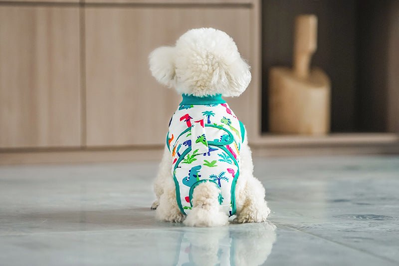 White dog wearing dinosaur print recovery suit with green trim, viewed from behind