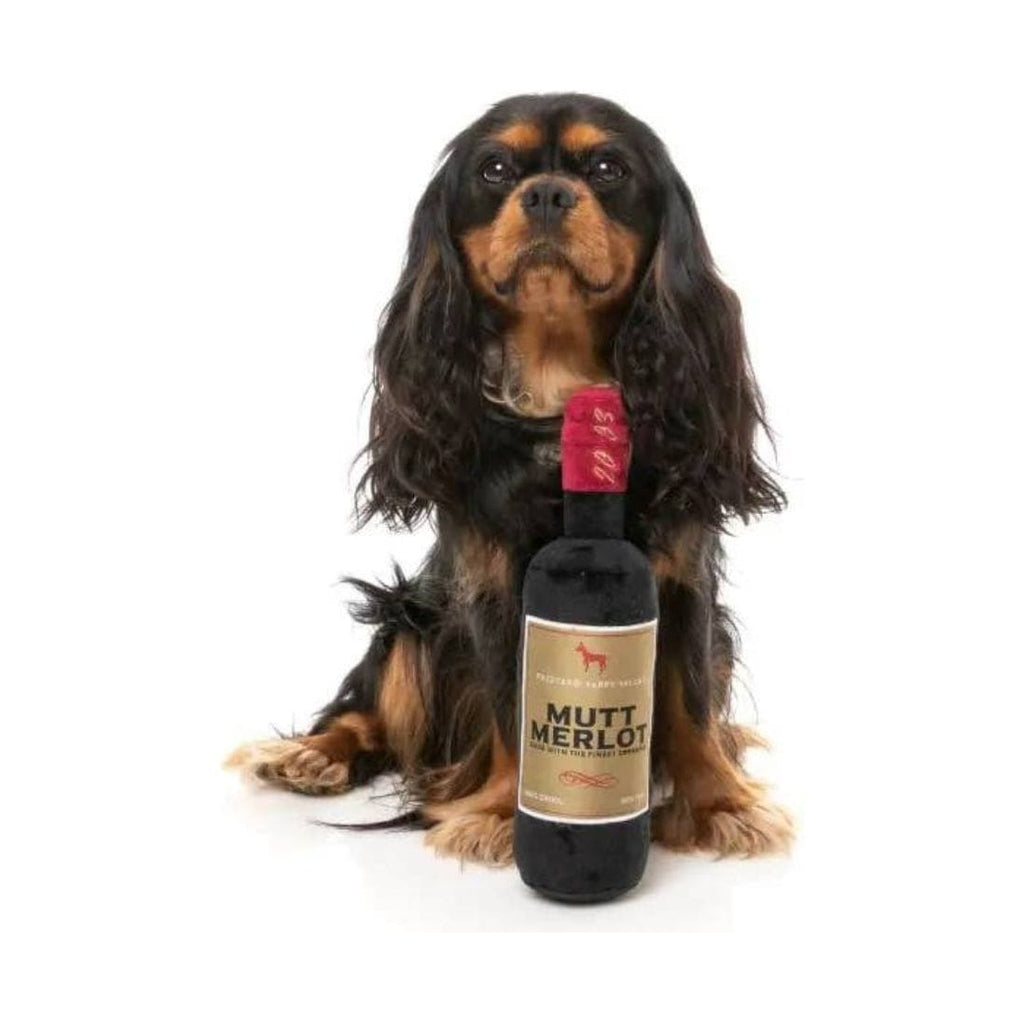 Plush dog toy bottle with MUTT MERLOT label, beside dark-and-tan dog